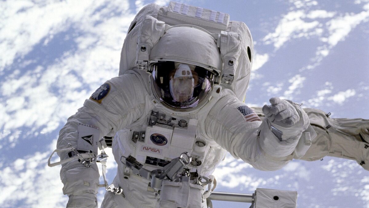 Astronaute en combinaison spatiale blanche flottant lors d'une sortie extravéhiculaire, visière reflétant l'intérieur d'un véhicule spatial, drapeau américain et logo NASA sur la combinaison, Terre et nuages bleus en arrière-plan.