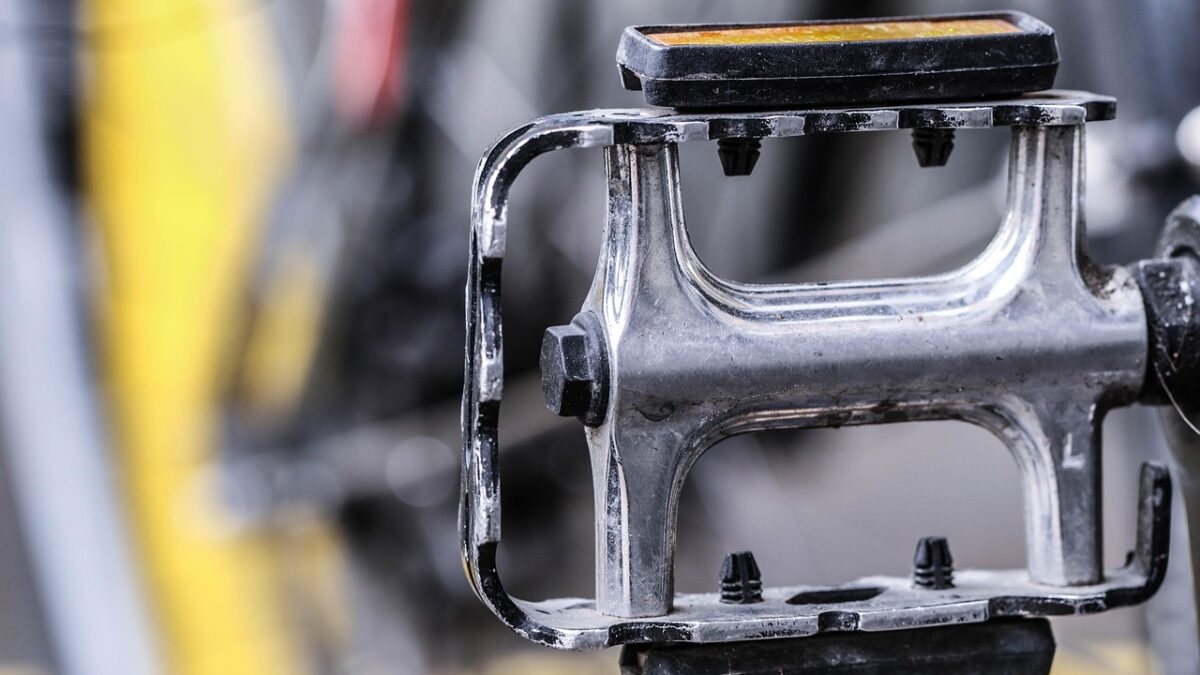 Gros plan sur une pédale de vélo plate en métal chromé, avec un catadioptre noir et jaune sur le dessus et des picots antidérapants, sur fond flou jaune et gris.