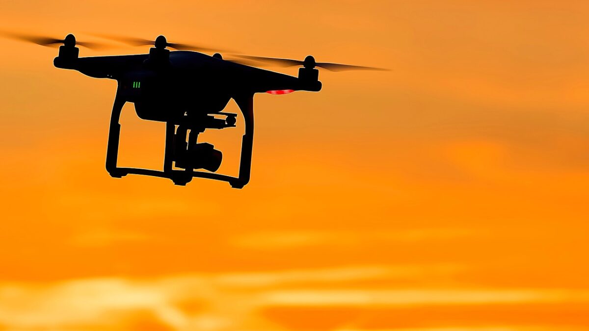 Silhouette d'un drone quadricoptère équipé d'une caméra sous la coque, en vol devant un ciel orange au coucher du soleil.