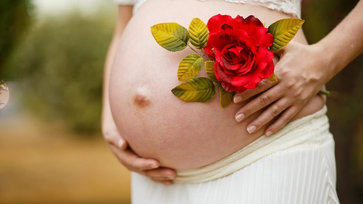 Gros plan sur le ventre d'une femme enceinte vêtue de blanc, orné d'une rose artificielle rouge et de feuilles, mains posées dessus, sur fond de verdure floue.