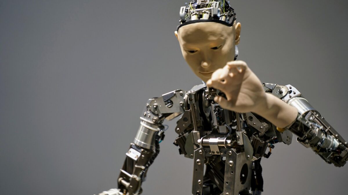 Robot humanoïde aux bras et au torse métalliques, à la tête beige et au visage lisse, tendant la main vers une main humaine au premier plan, sur fond gris uni.