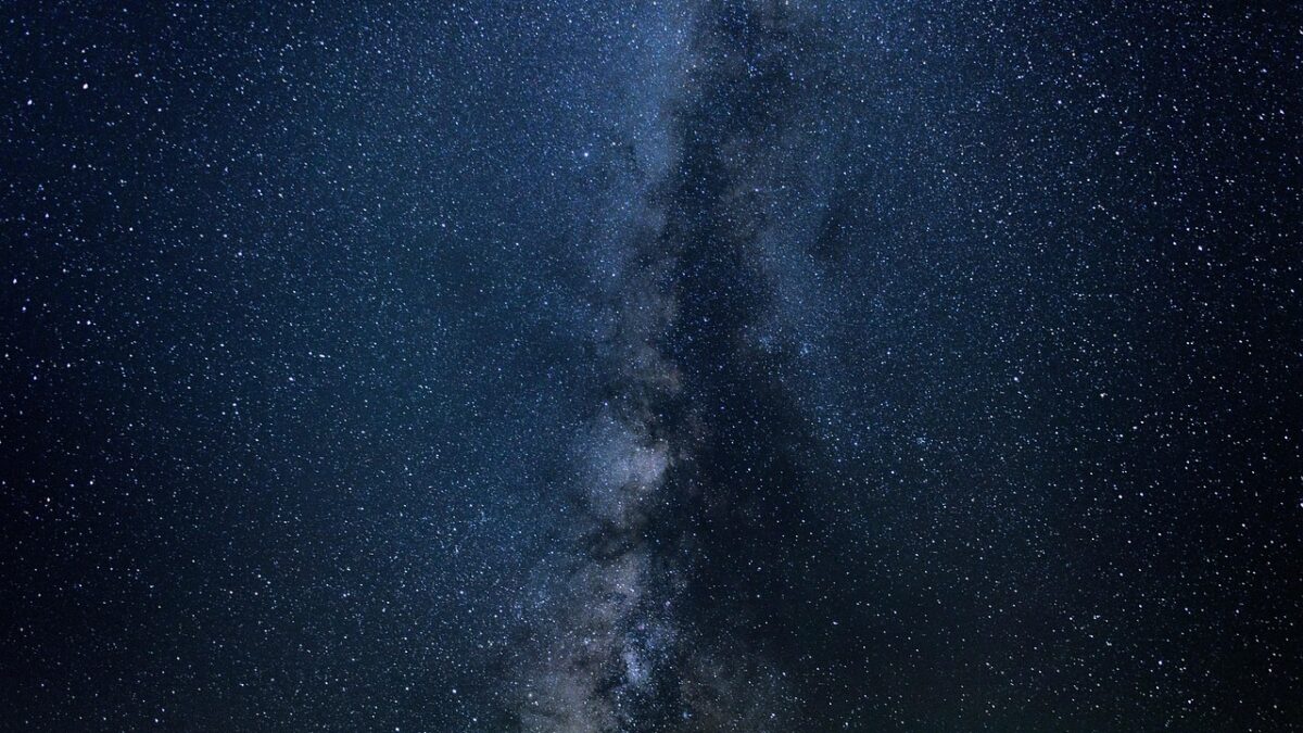 Ciel nocturne étoilé traversé verticalement par la bande de la Voie lactée, avec des zones sombres de poussière et des nuages lumineux.