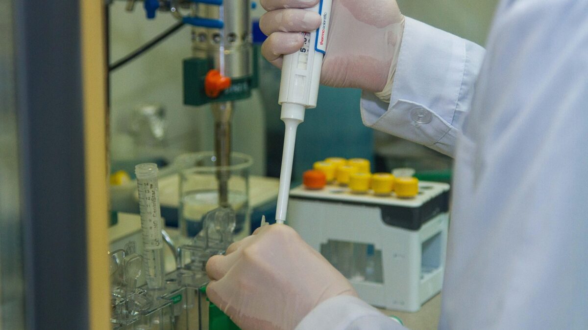 Gros plan sur les mains gantées d'un chercheur en blouse blanche utilisant une micropipette blanche au-dessus d'un tube à essai et de tubes à capuchons jaunes dans un portoir, sur fond d'appareillage de laboratoire.