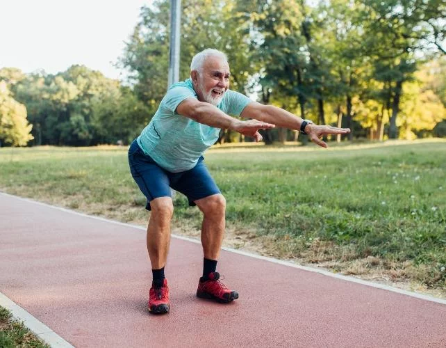 Un homme plus âgé accroupi, bras tendus, dans un parc