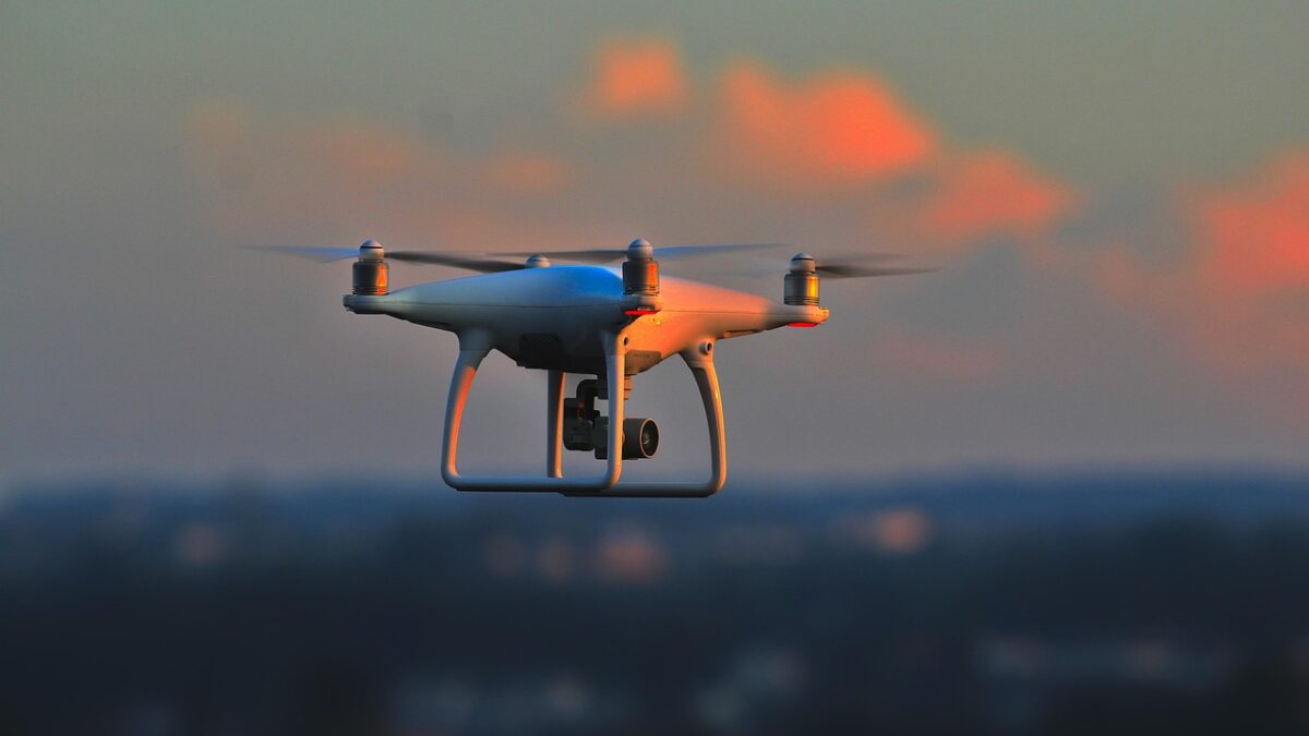 Un drone quadricoptère blanc en vol au coucher du soleil, caméra fixée sous le châssis, sur fond de ciel orangé et de ville floutée.