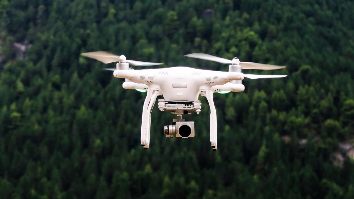 Un drone quadricoptère blanc en vol stationnaire, caméra fixée sous le fuselage, au-dessus d'une forêt de conifères verte.