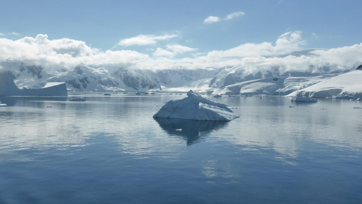 Mer calme et bleutée, icebergs flottants et montagnes enneigées sous un ciel nuageux en Antarctique.