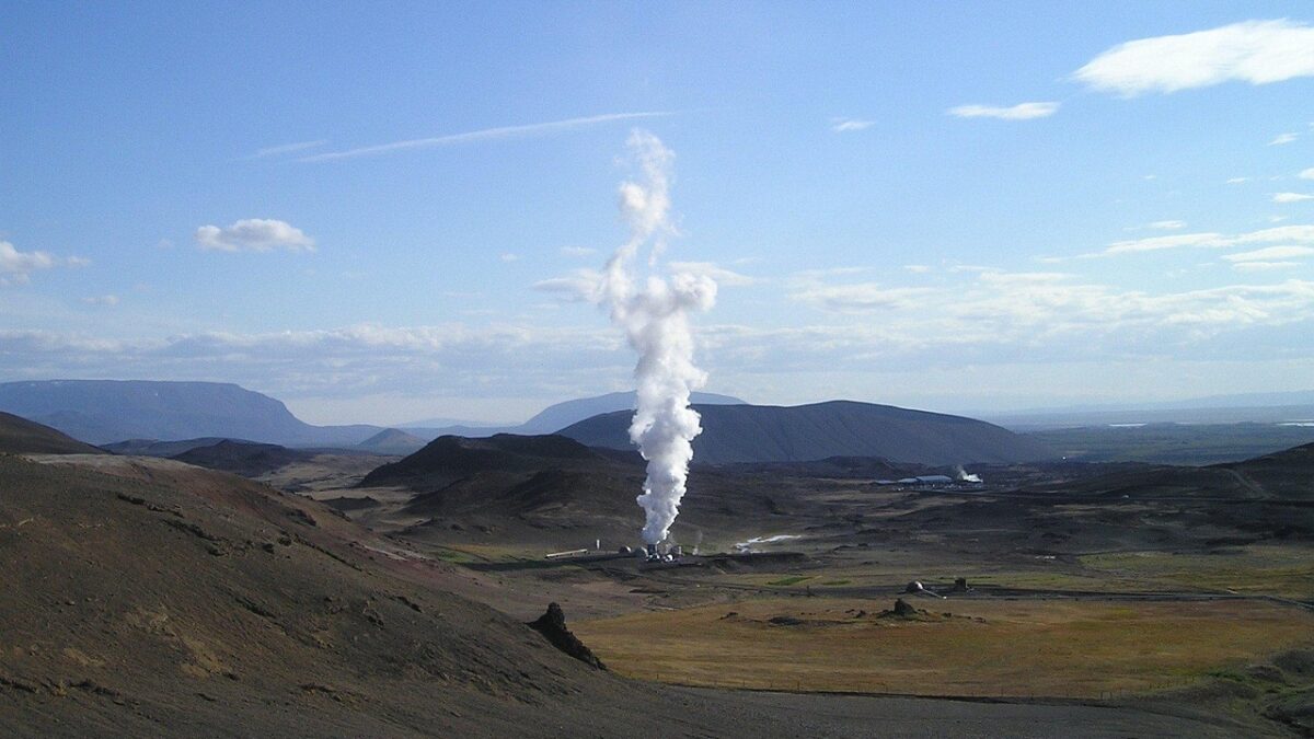 Paysage volcanique aride avec une centrale géothermique au centre, d'où s'échappe un panache de vapeur blanche sous un ciel bleu partiellement nuageux.