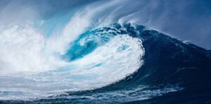 Une grosse vague bleue se courbe et déferle en écume blanche sous un ciel nuageux, vue en mer.