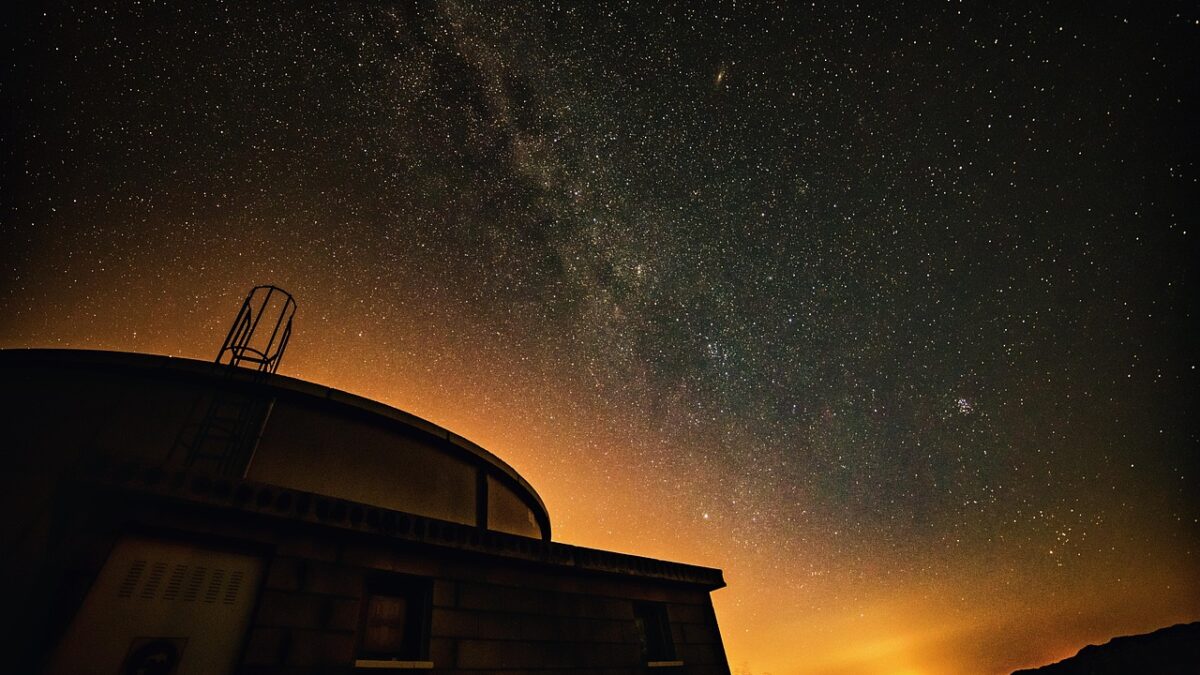 Coupole d'observatoire dans un bâtiment sombre, sous un ciel nocturne étoilé avec la Voie lactée et une lueur orangée à l'horizon.