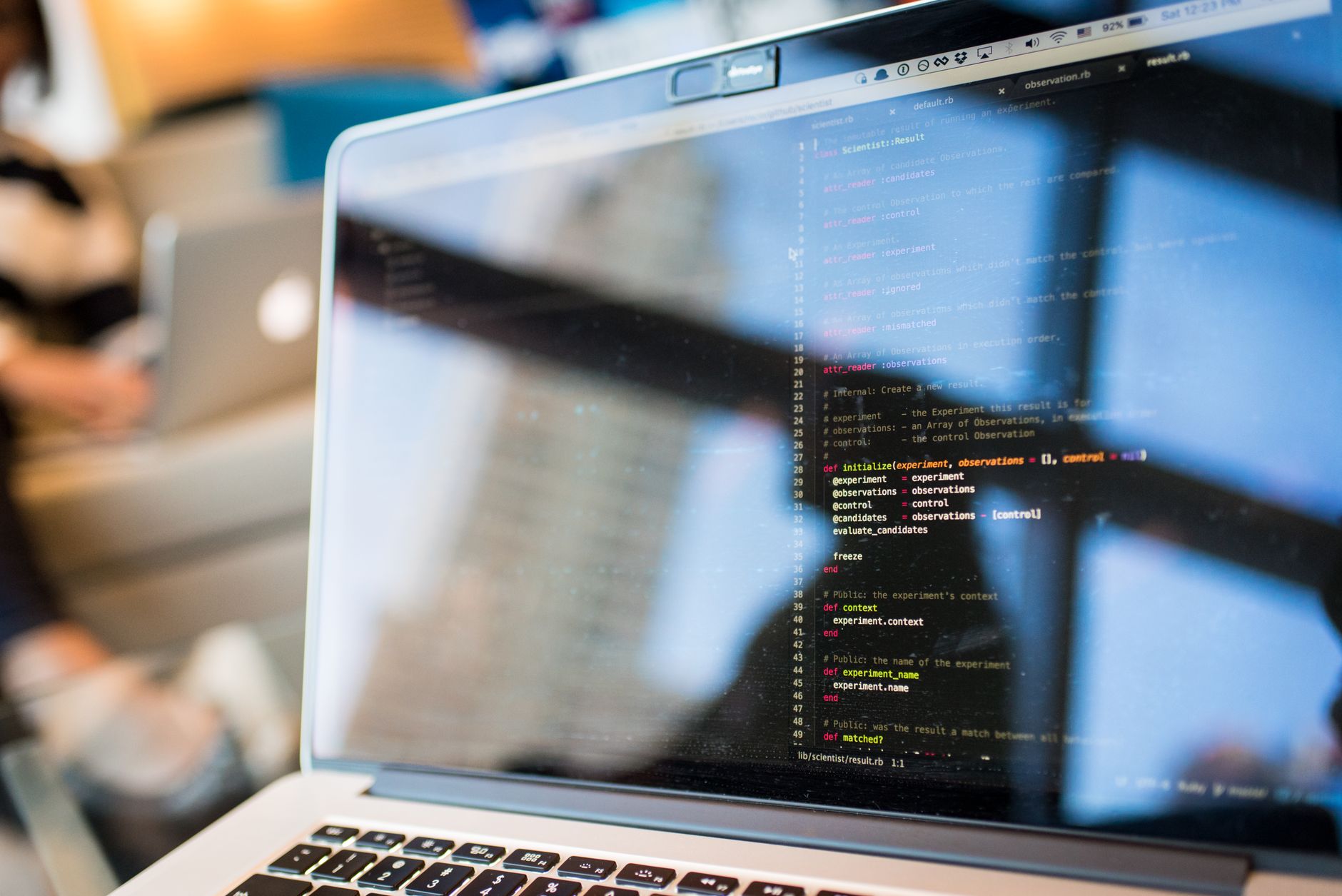 Écran d’ordinateur portable affichant du code informatique, avec des reflets de fenêtres et une personne floue à l’arrière-plan.