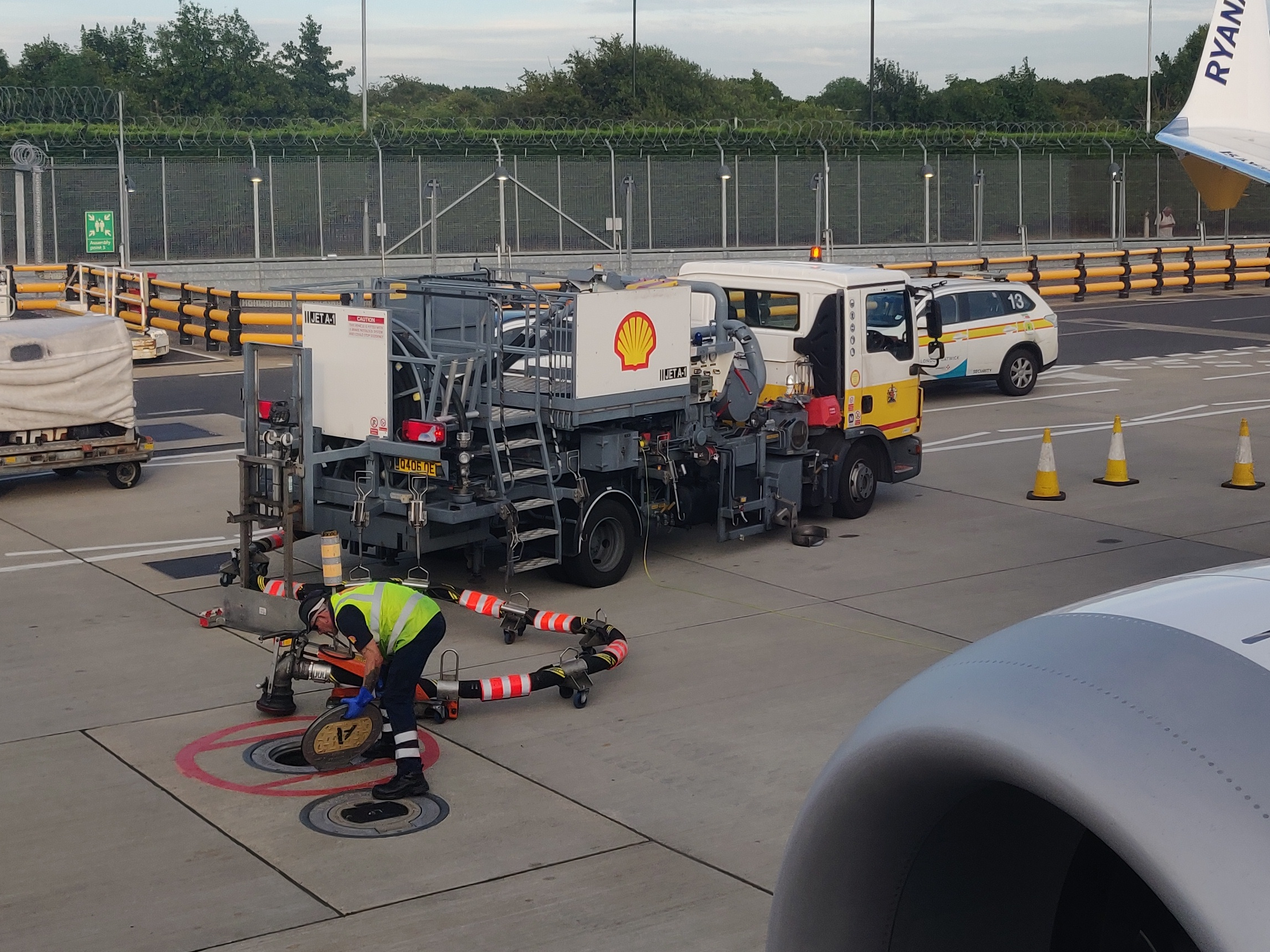 Un agent en gilet jaune ouvre une bouche de ravitaillement près d’un camion avitailleur Shell sur l’aire d’un aéroport, à côté d’un moteur d’avion.