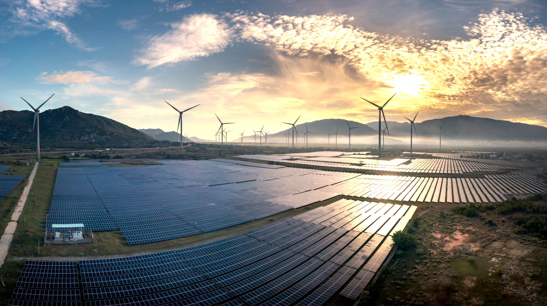 Vue aérienne de panneaux solaires et d’éoliennes au pied de montagnes, sous un ciel nuageux au soleil bas.